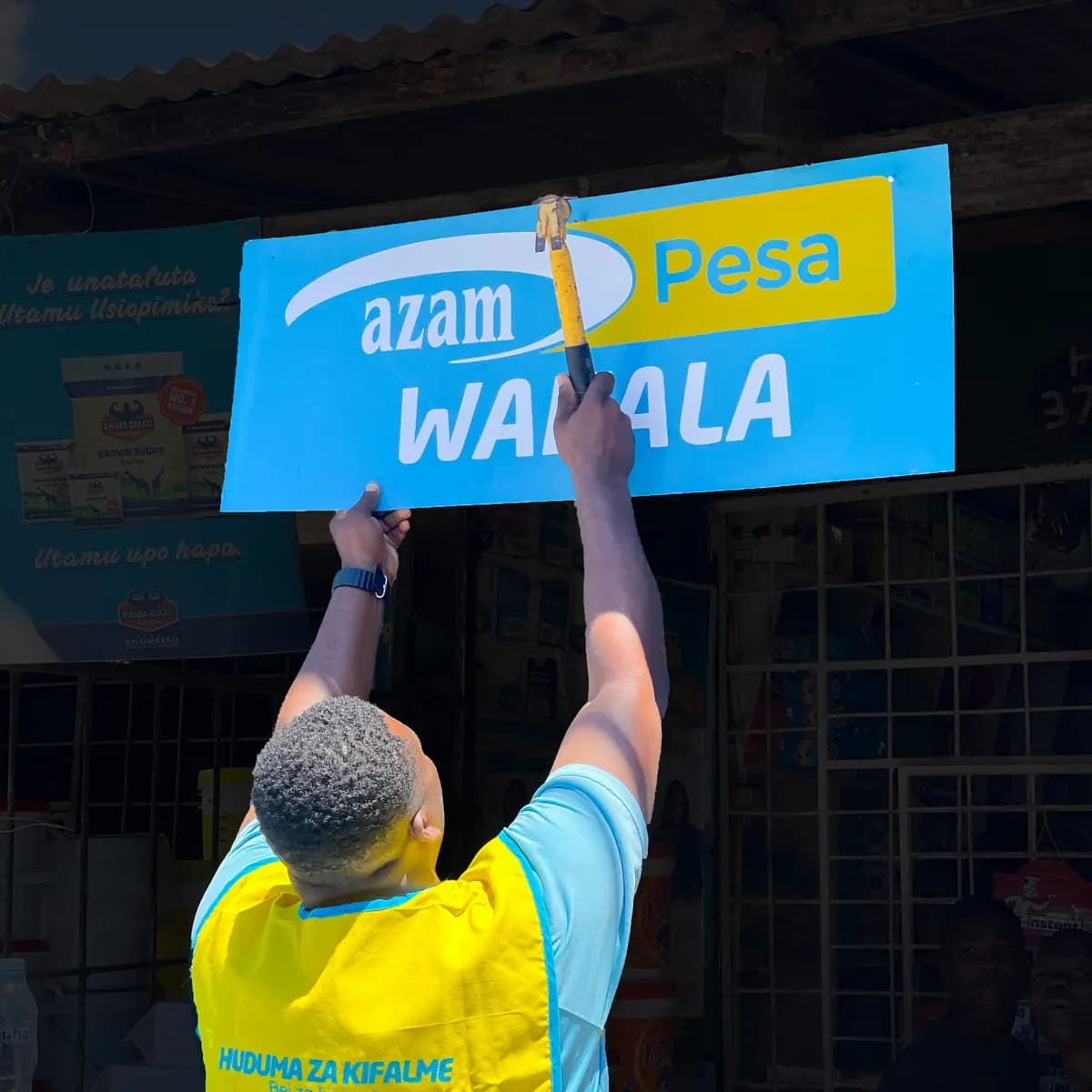 Wakala akiweka bango la AzamPesa Wakala juu ya duka lake